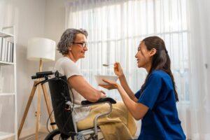 Asiatische Pflegerin, Krankenschwester, reicht einer älteren Frau zuhause das Essen an. Fachpflegekraft unterstützt und kümmert sich um ältere Frau im Wohnzimmer.